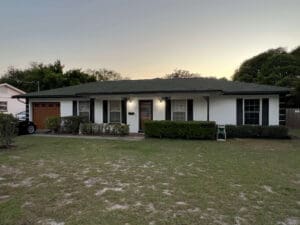 A single-story white house with a dark roof, garage, and well-maintained shrubs at sunset. A car is parked in the driveway, with trees and a patchy lawn in the foreground.
