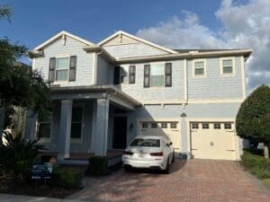 A two-story light gray house with a brick driveway and a white car parked in front under a blue sky with clouds.