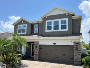 A two-story house with a gray exterior, white trim, and a brick driveway under a blue sky with clouds. The front features a garage, lush greenery,