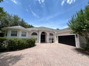 A single-story house with a light pink exterior, dark roof, and a brick driveway under a blue sky.