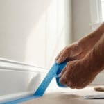 Professional painter applying blue painter's tape along baseboard trim during interior wall preparation.